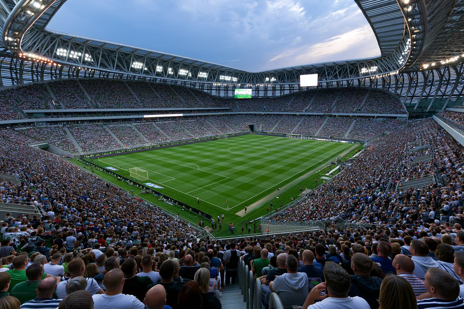 Ein großes Fußballstadion, gefüllt mit Fans unter Flutlicht an einem Sommerabend.