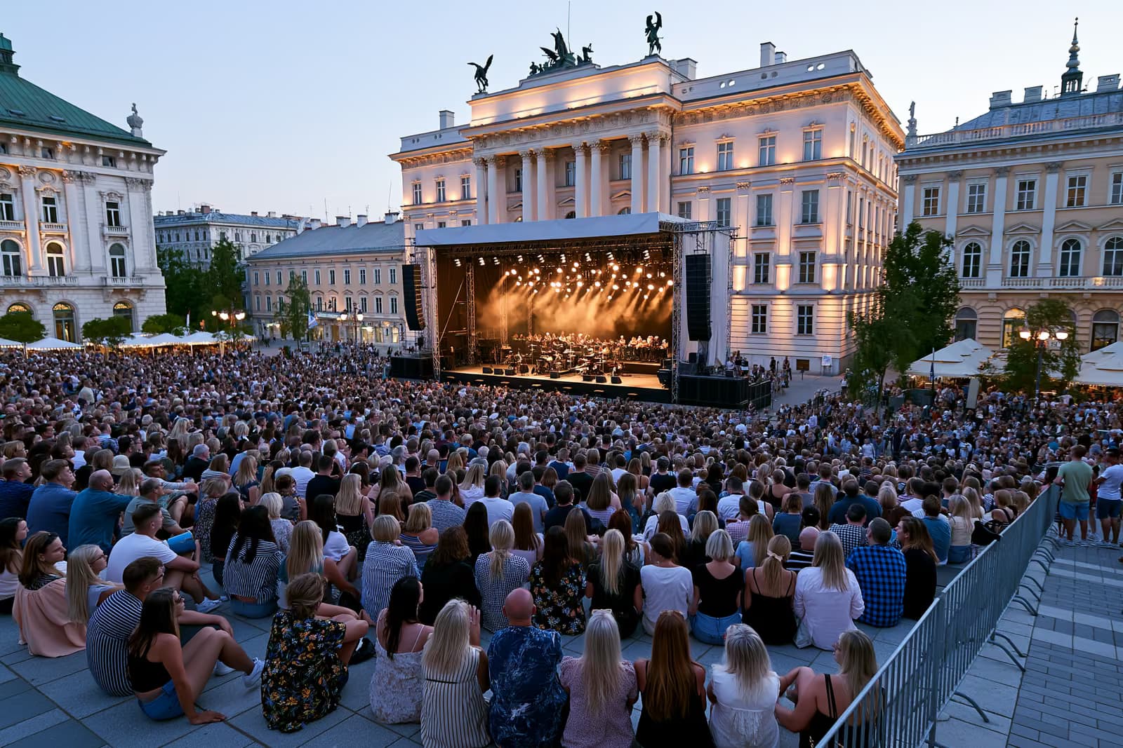In der Abenddämmerung versammelt sich eine Menschenmenge auf einem historischen Berliner Platz, wo eine Freilichtbühne vor prächtigen Fassaden beleuchtet wird.