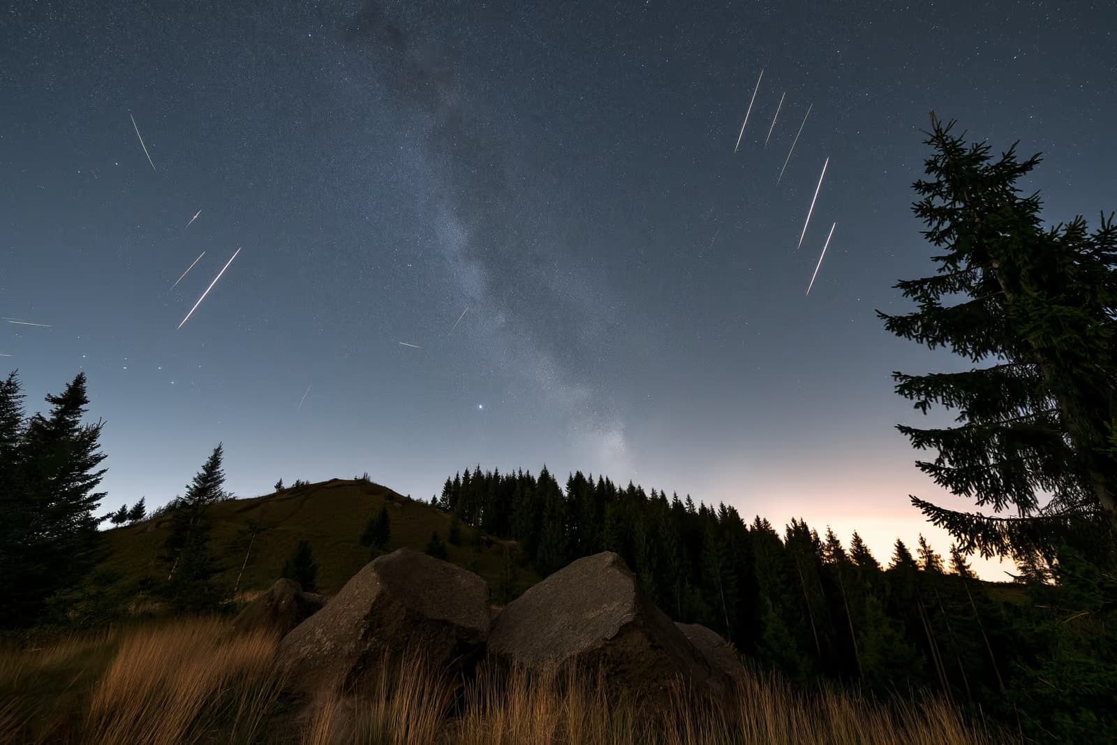Ein Nachthimmel über einer dunklen deutschen Landschaft mit Meteoritenspuren und einem schwachen Sternenband darüber.