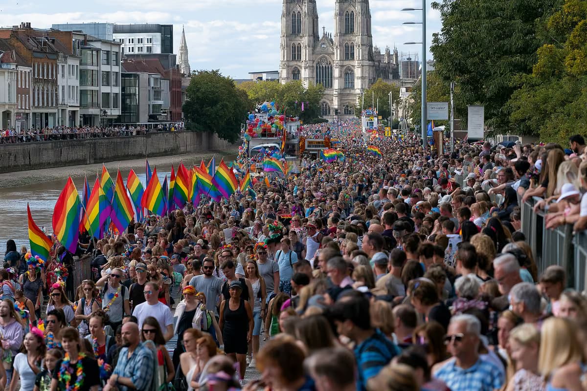 Regenbogenfahnen und geschmückte Festwagen ziehen durch eine belebte Straße am Flussufer.