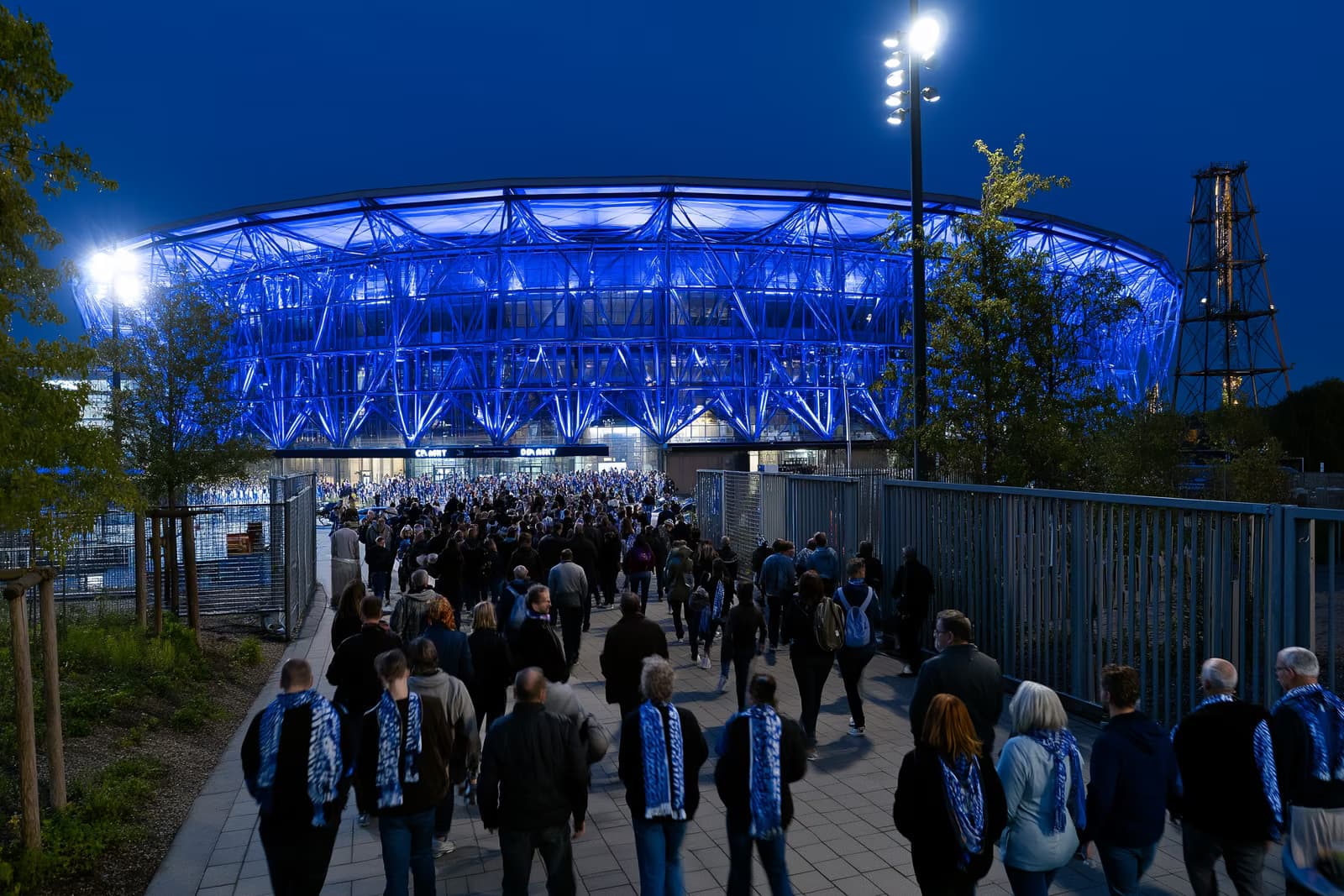 Ein Fußballstadion im Ruhrgebiet erstrahlt nachts in Königsblau, während Fans mit Schals nach dem Wechsel von Gosens zu Schalke zu den Toren strömen.